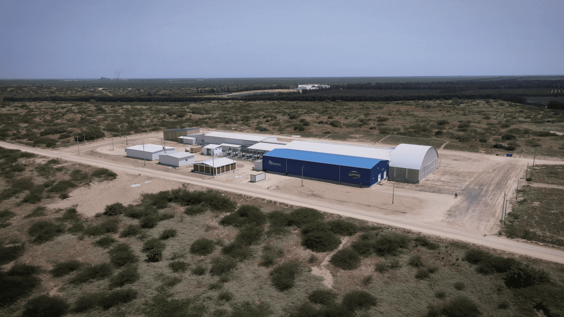 Vista aérea de una instalación industrial con varios edificios y almacenes rodeados por un camino de tierra y escasa vegetación.